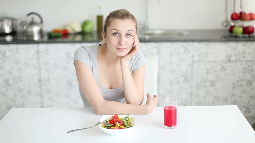 Beautiful young woman sitting at kitchen table with bowl of salad and glass of