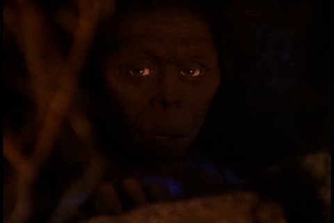 Historical reenactment in East Africa. Early human, or Homo erectus, peers out from behind rock. Fire light flickers across her face.