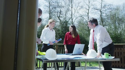 Business group come together for an outdoor meeting on a bright sunny day. They are on a terrace looking at construction plans, could be a team of architects, engineers or designers. In slow motion.