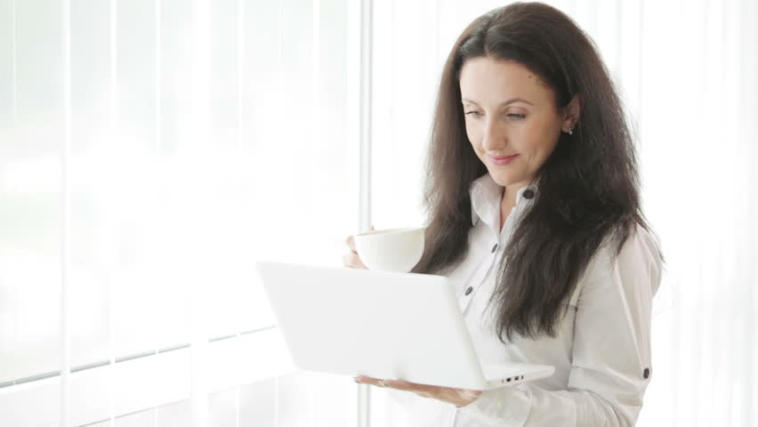 Beautiful young woman standing in front of window using laptop drinking coffee