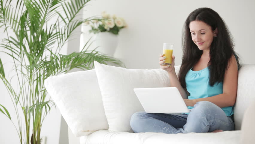 Pretty brunette woman sitting on sofa using laptop drinking juice and smiling at
