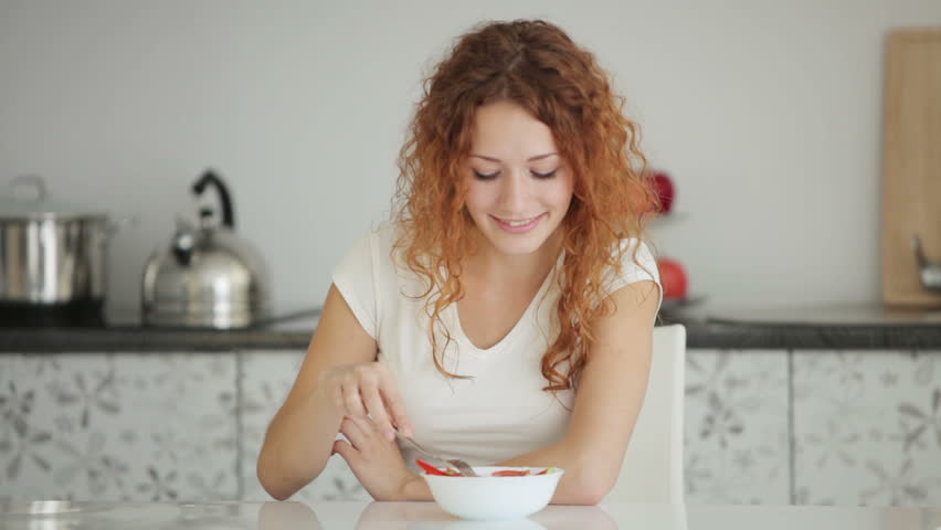 Cute young woman sitting at table in kitchen eating salad and smiling