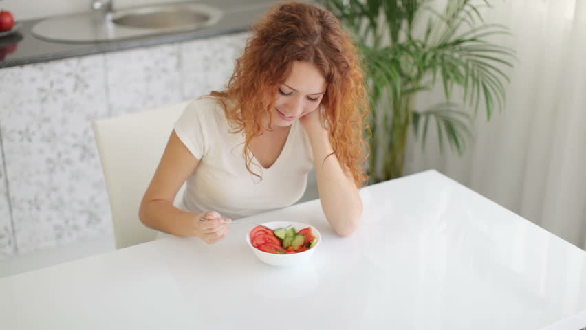 Attractive young woman sitting at kitchen table and eating vegetable salad with