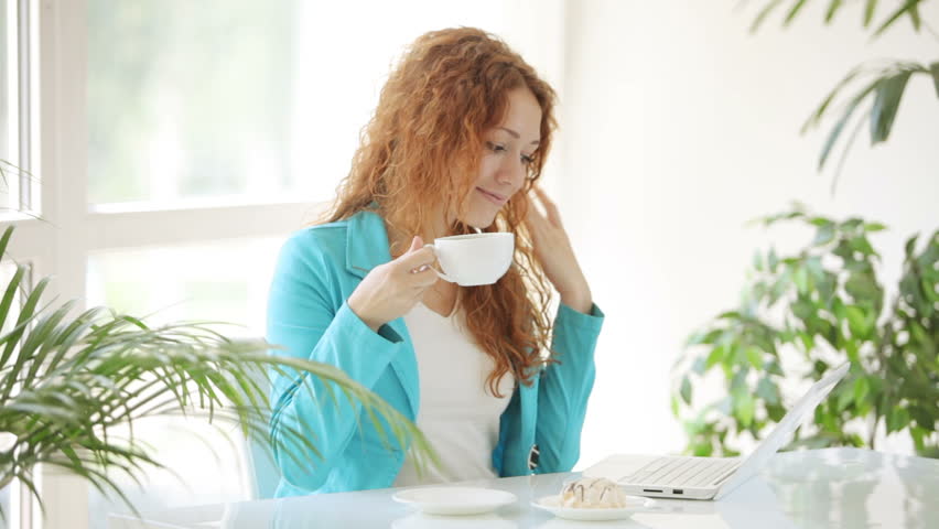 Attractive young woman sitting at table using laptop and drinking coffee with