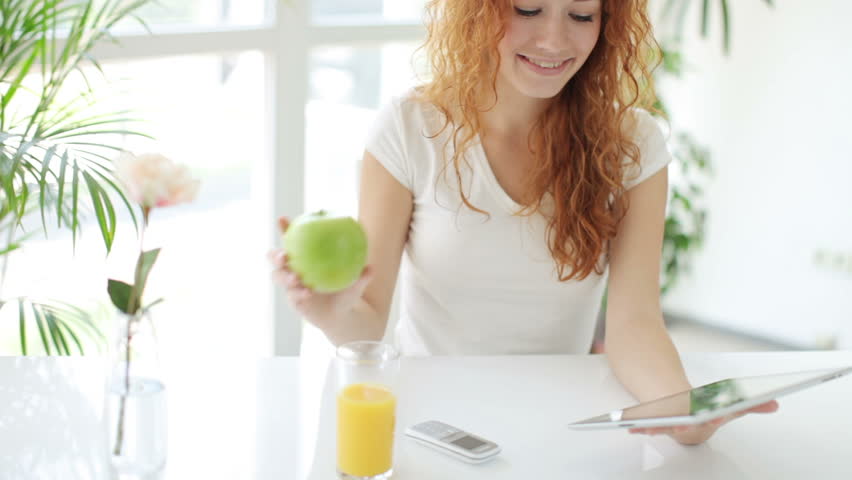 Attractive young woman sitting at table eating apple using touchpad and smiling