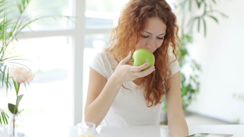 Pretty girl sitting at table using touchpad eating apple and smiling