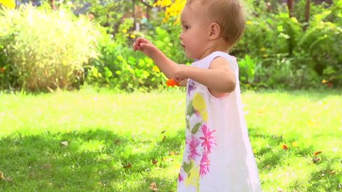 Adorable one year old Baby Girl making her first steps outdoor. Slow Motion Video Footage of the First Steps of the Kid. Sunny day and Green Grass 