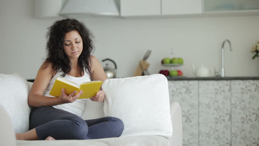 Pretty young woman sitting on sofa reading book closing it and smiling at camera