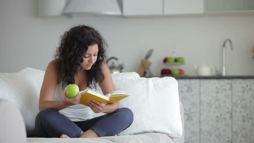 Pretty young woman sitting on sofa with book eating apple and smiling