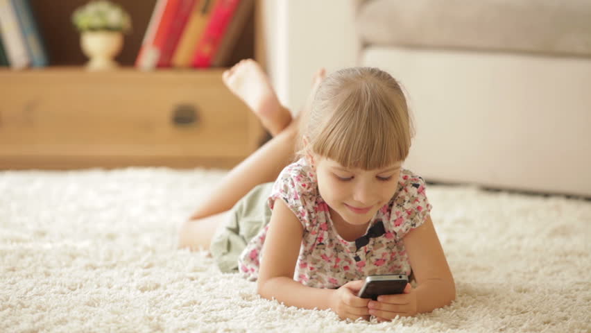Cheerful little girl lying on carpet using mobile phone and smiling at camera