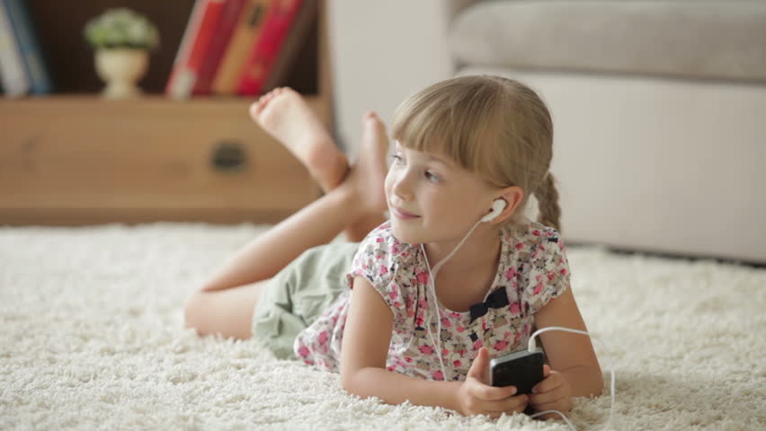 Cute little girl lying on floor listening to music on mp3 player and smiling at
