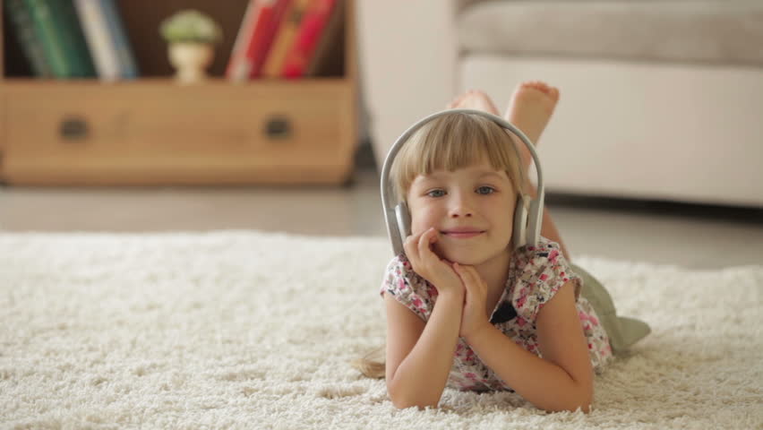 Cheerful little girl lying on carpet in living room listening to music on