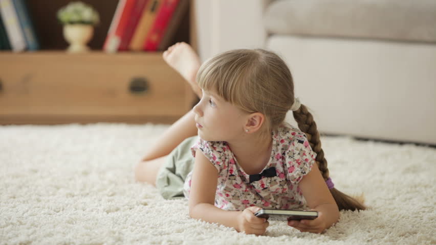Beautiful little girl lying on floor using cellphone and smiling at camera