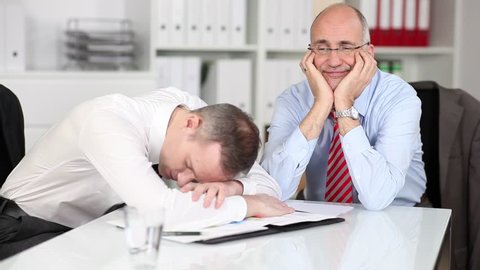 Two despondent tired businessmen sitting together at a table with the younger man collapsed over the paperwork and his colleagues staring up into the air with a weary expression