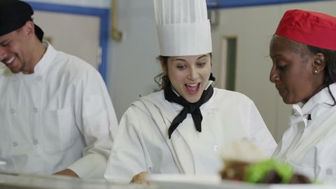 Mixed ethnicity team of professional chefs preparing and cooking food in a commercial kitchen. The head chef tastes a dish and gives her approval. In slow motion