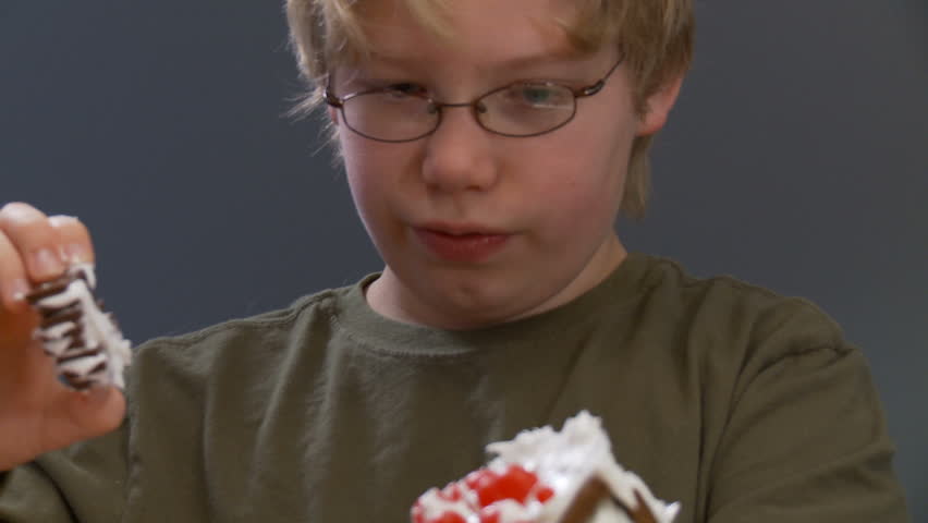 Boy sticks a chimney on a gingerbread house then licks his fingers.