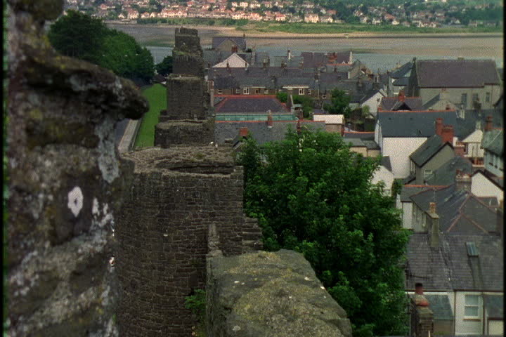 High stone wall bordering village, camera pans right over village rooftops and zooms in to Conwy Castle in North Wales.