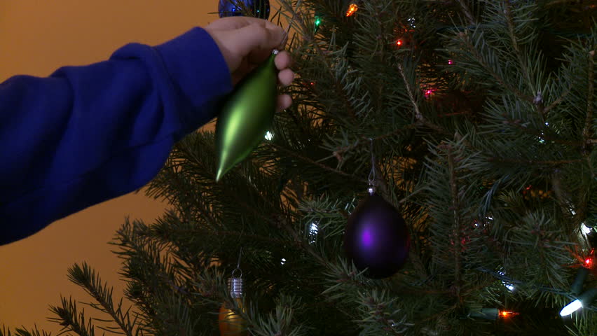Child's hand hangs a green ornament on a Christmas tree.