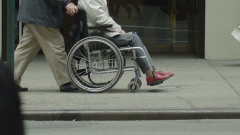 Low angle view of an elderly lady in a wheelchair being pushed along a crowded city street. In slow motion.