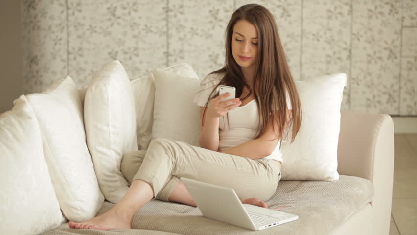Beautiful girl relaxing on sofa with laptop using mobile phone and smiling at