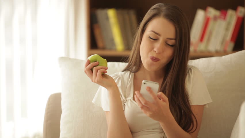 Beautiful girl sitting on sofa eating apple using cellphone and smiling