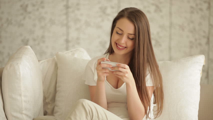 Attractive young woman sitting on sofa using cellphone and smiling