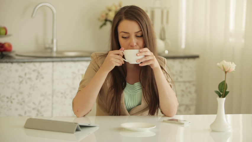 Beautiful girl sitting at table using touchpad drinking tea and smiling at