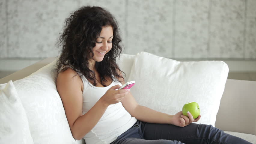 Beautiful young woman sitting on couch using mobile phone eating apple and