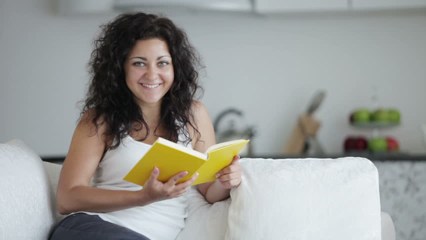 Beautiful young woman sitting on sofa reading book and smiling at camera