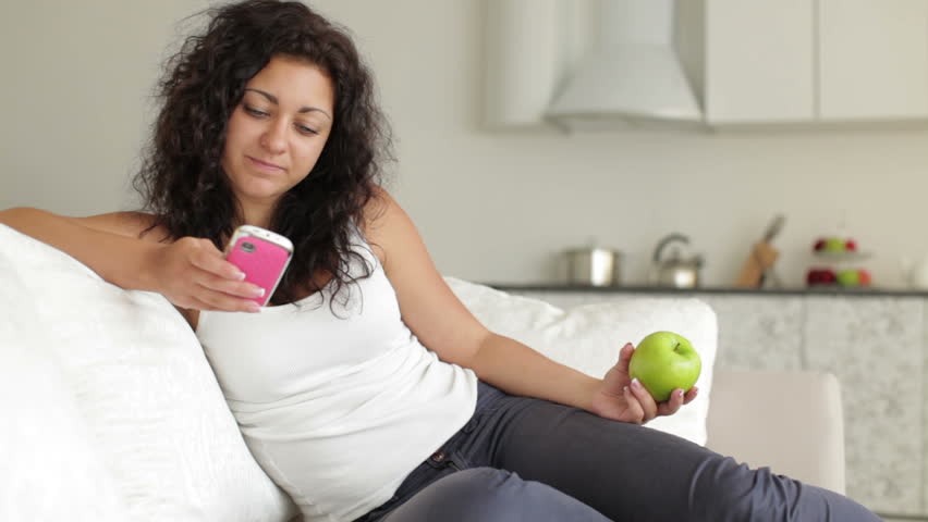 Cheerful young woman relaxing on sofa using mobile phone eating apple and