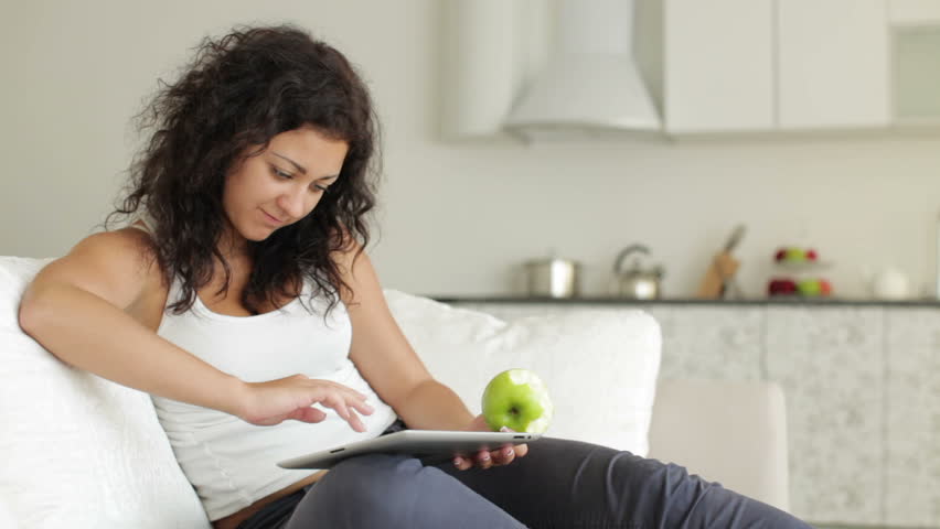 Charming young woman sitting on couch using touchpad eating apple and smiling