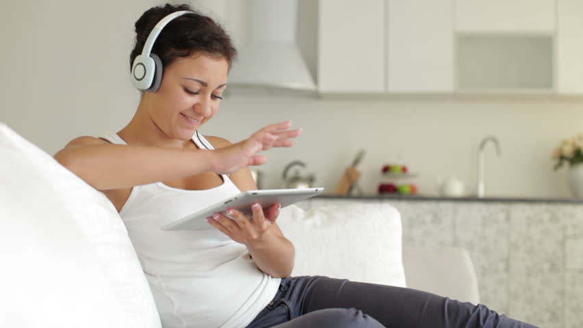 Attractive young woman in headphones sitting on sofa smiling and using touchpad