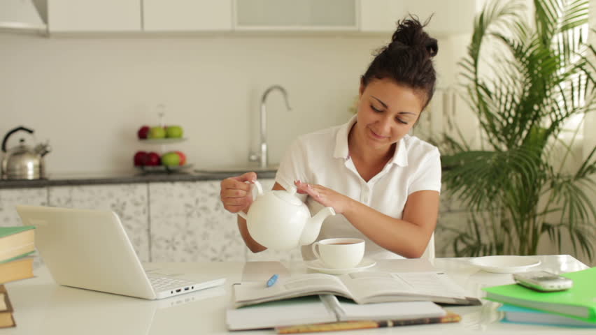 Cheerful young woman sitting at table with books and laptop pouring tea into cup