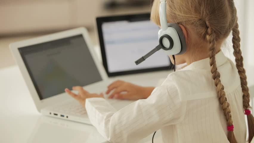 Cheerful little girl in headset with microphone sitting at table using laptop