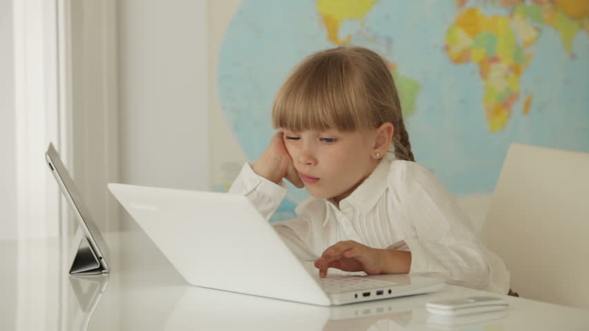 Bored little girl sitting at desk using touchpad while studying with laptop
