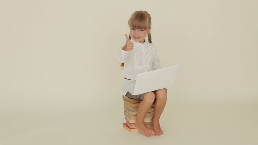 Beautiful little girl sitting on pile of books with laptop showing thumb up and