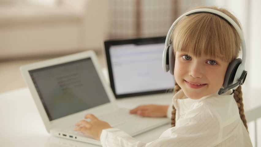 Beautiful little girl in headset sitting at table working on laptop turning