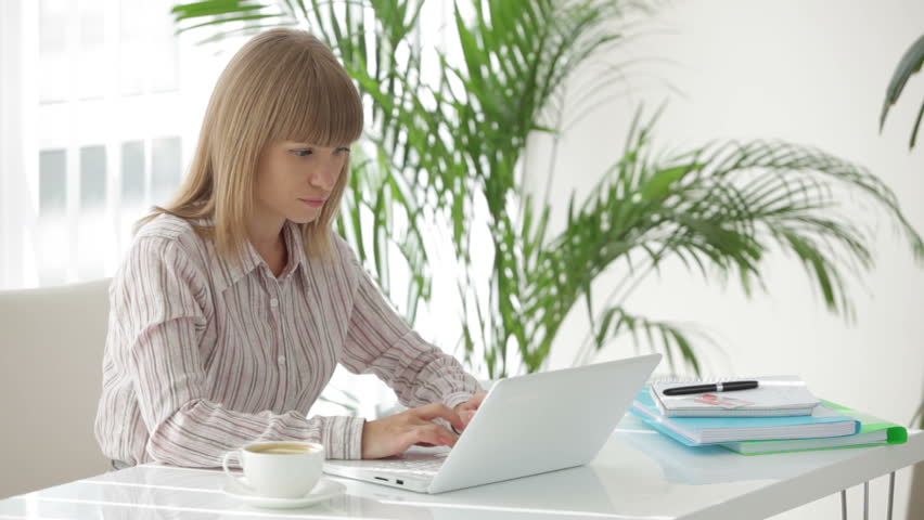 Young businesswoman sitting at table using laptop holding cup of coffee in her