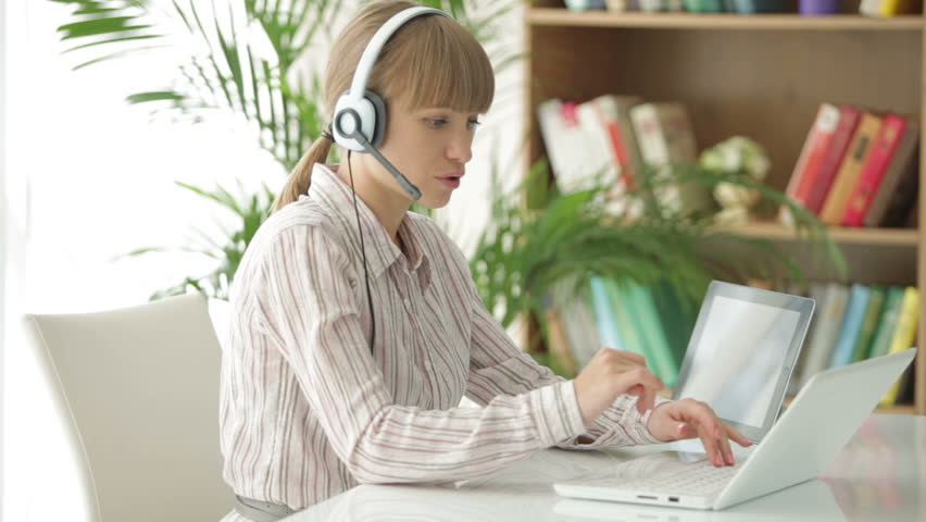 Pretty young woman in headset sitting at table using laptop and touchpad and