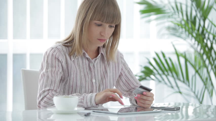 Smiling young woman sitting at table using touchpad and holding credit card in