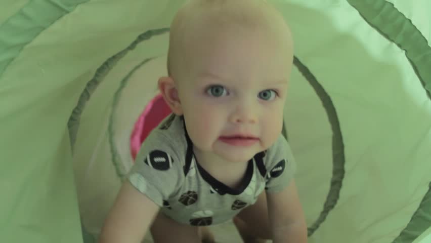 A baby boy playing with a green tunnel in his house.