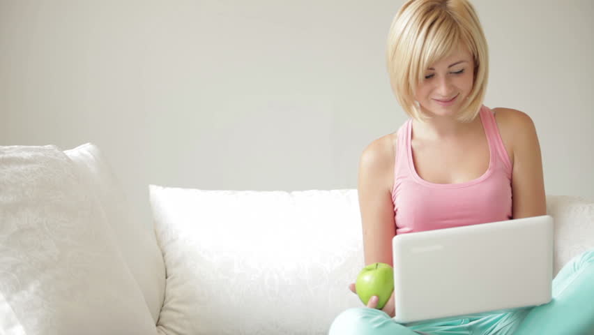 Cute smiling girl sitting on sofa eating green apple and using laptop