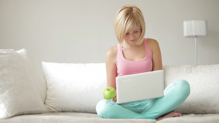 Smiling girl sitting on couch with crossed legs eating apple and using laptop