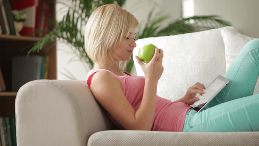 Pretty young woman relaxing on couch eating apple using touchpad and smiling