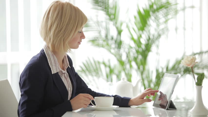Pretty young woman sitting at office table using laptop and pouring tea into cup