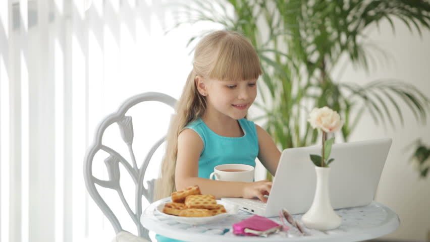 Funny little girl sitting at table with plate of biscuits and cup of tea using