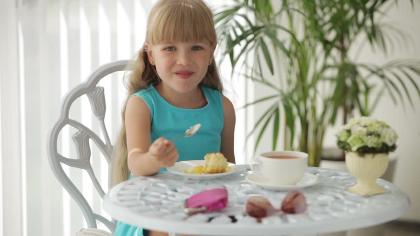 Smiling little girl sitting at table drinking tea and eating cake