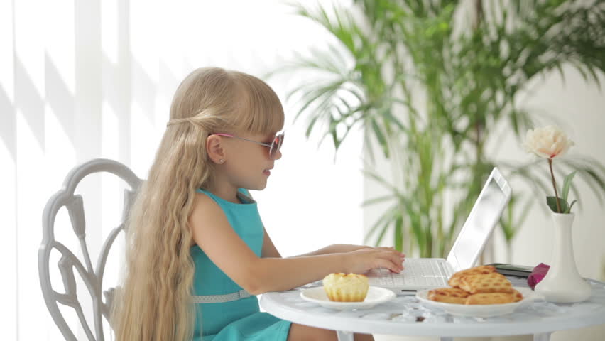 Funny little girl wearing sunglasses sitting at table using laptop and eating
