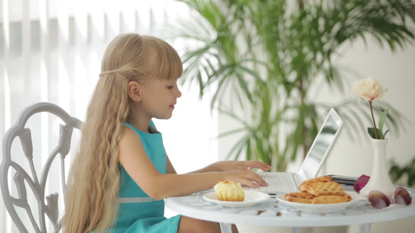 Pretty little girl sitting at table eating cake using laptop and smiling at