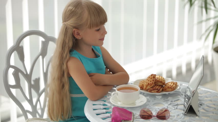 Little girl sitting at table with plate of biscuits and cup of tea using